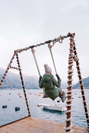 Young woman swings on a swing on the pier and looks at the mountains. Side view
