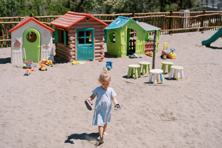 Little girl walks barefoot on the sand on the playground with sandals in her hands