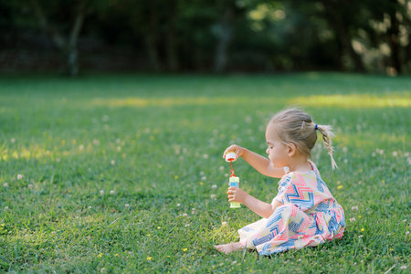 Little girl is sitting on a green meadow with a bottle of soap bubbles in her hands. Side view