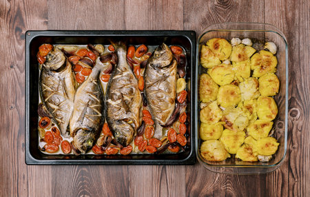 Baked sea bream with vegetables on a baking sheet stands with baked potatoes in a dish on a wooden table. Top viewの写真素材