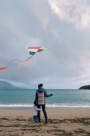 Small girl hugs her mother leg standing on the seashore with a kite. Back viewの写真素材