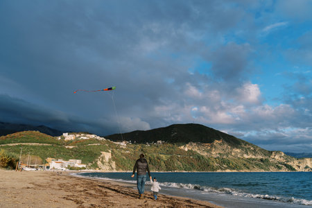 Dad and little daughter are walking along the seashore with a soaring kite. Back viewの写真素材