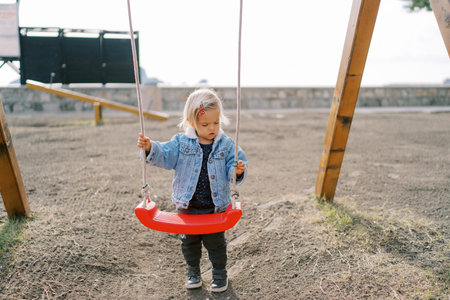 Little girl stands near the swing on the playground holding on to the ropesの写真素材