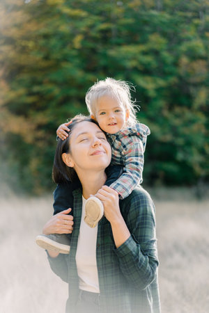 Smiling little girl hugging her mother head while sitting on her shouldersの写真素材