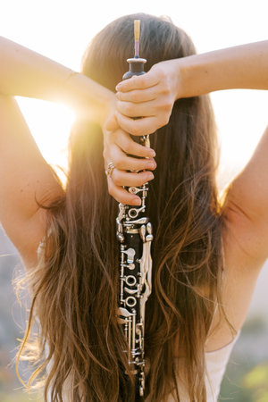 Girl with a clarinet in her hands behind her head stands in the sun. Back viewの写真素材