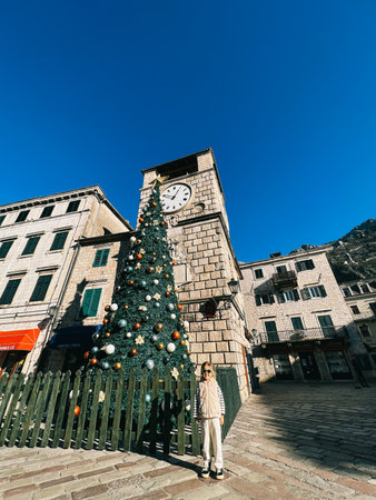 Little girl stands near a Christmas tree in front of an ancient building with a clockの写真素材