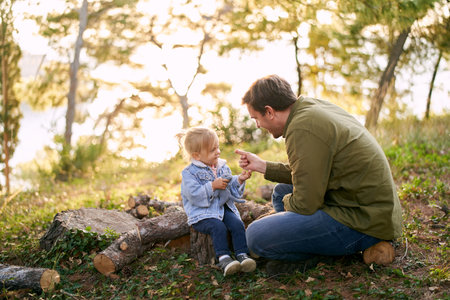 Dad holds out a blade of grass to a little smiling girl sitting on logs in the forestの写真素材