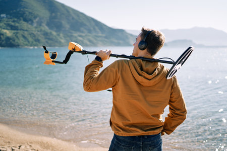 Beach cop stands with a metal detector on his shoulder on the shore and looks into the distance. Back viewの写真素材