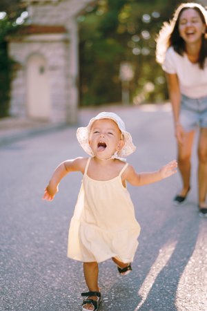 Little girl runs along the road in the park with her mouth open against the background of a laughing motherの写真素材