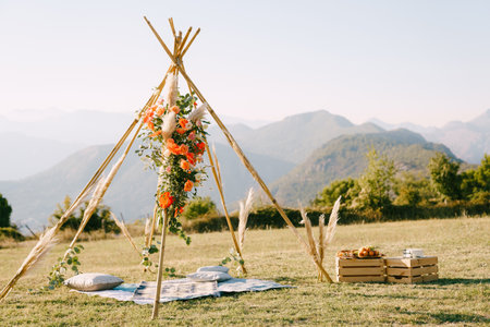 Boho-style wedding arch stands near a bedspread with pillows and boxes of food in a clearingの写真素材