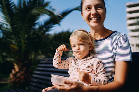 Little girl eats porridge with a spoon while sitting on the lap of a laughing motherの写真素材