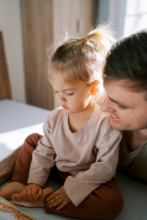 Little girl is looking intently at the book that her dad is reading to her while sitting on the bedの写真素材