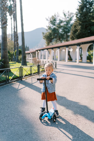 Little girl stands near a scooter holding the steering wheel on the road in a green parkの写真素材