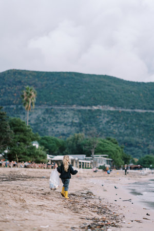 Little girl with a bag walks along a dirty beach collecting trash. Back viewの写真素材