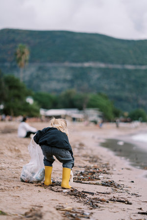 Little girl collects garbage in a bag while crouching on the beach. Back viewの写真素材
