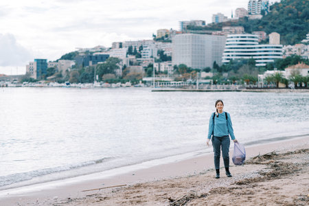 Young woman with a bag of garbage wearing gloves stands on a sandy beach near the seaの写真素材