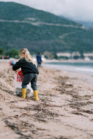 Little girl puts a bottle in a bag of trash on a sandy beach. Back viewの写真素材