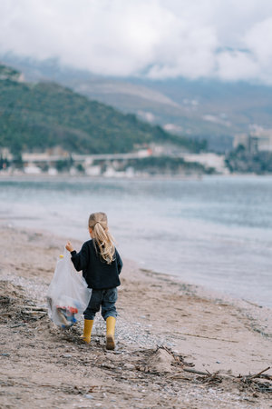 Little girl with a bag of garbage walks along the beach along the sea. Back viewの写真素材
