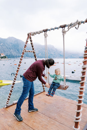 Mom swings a little girl on a swing on a pier by the sea