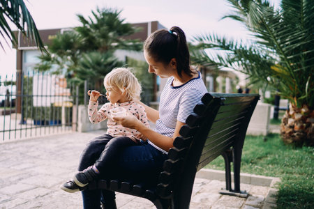 Little girl eats porridge with a spoon from a box in the hands of her mom sitting on a park benchの写真素材