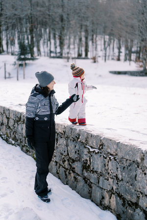 Little girl walks along a fence in a winter park, holding her mother hand and pointing to the sideの写真素材