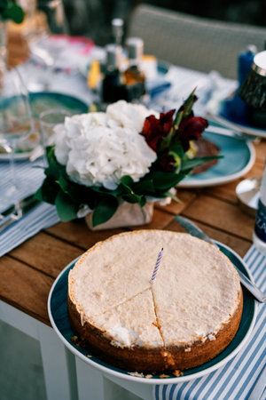 Cake with a cut out sector and a candle stands on a plate near a bouquet of flowers on the table