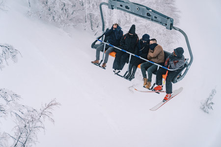 Skiers in ski equipment go uphill on a chairlift above snow-covered treesの写真素材
