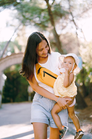 Mom with a little girl in her arms, drinking water from a bottle, walks along the road in the park