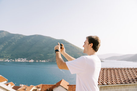 Young man takes a photo with a smartphone of a view of the mountains and the sea over the red roofs of housesの写真素材