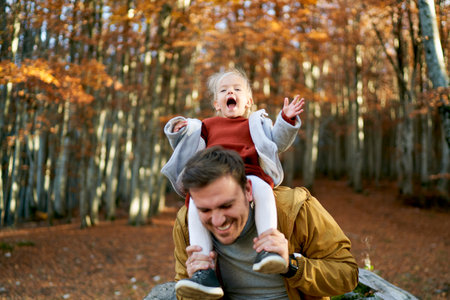 Little laughing girl waves her hands while sitting on the shoulders of her dad, who leaned forwardの写真素材