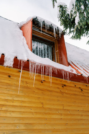 Row of icicles hangs from the snow-covered roof and attic window of a wooden cottageの写真素材