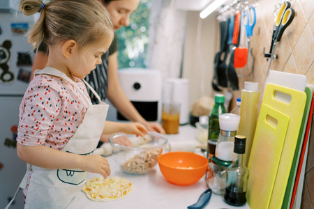 Little girl puts toppings on rolled out pizza dough while standing with her mom at the kitchen tableの写真素材