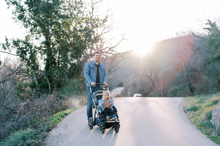 Smiling dad carries his little daughter in a stroller along the road from the mountain