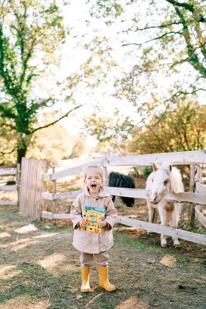Little girl with colorful book screams while standing in front of pony paddock on farmの写真素材
