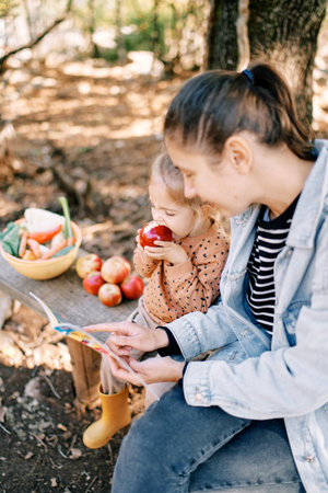 Mom reads a book to a little girl gnawing an apple on a bench in the gardenの写真素材