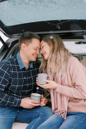 Smiling boyfriend and girlfriend touch foreheads while sitting with coffee mugs in car trunkの写真素材
