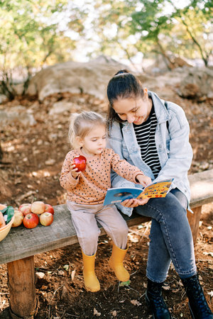 Little girl with an apple in her hand points to a book in her mother hands while sitting on a bench in the gardenの写真素材