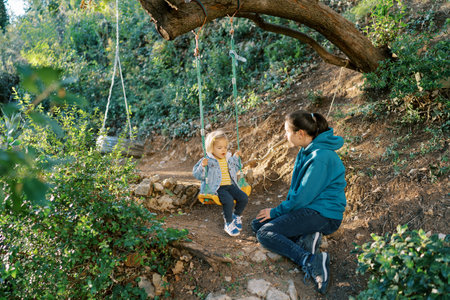 Mom swings a little girl on a rope swing sitting on the ground nearby