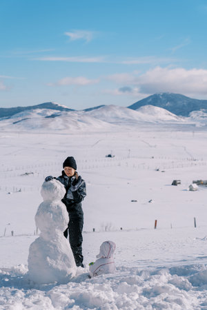 Small child sits in the snow and watches his mother build a snowman in a mountain valley. Back viewの写真素材