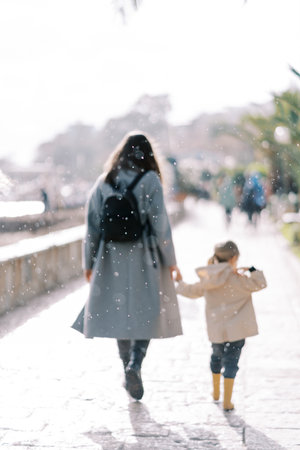 Mom and little girl are walking holding hands under the snowfall on the embankment. Back viewの写真素材