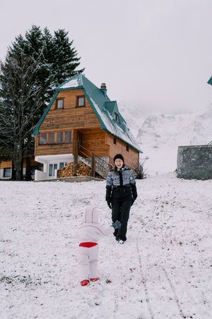Small child walks towards his laughing mother along a snow-covered hill near a wooden cottage. Back viewの写真素材