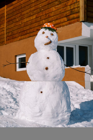 Snowman with a saucepan with red polka dots on his head stands near a wooden house