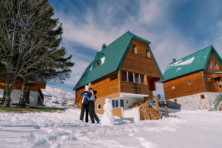 Mom kisses dad with little girl on his shoulders standing next to snowman near wooden chalet