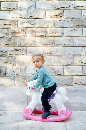 Little girl sitting on a toy unicorn rocking chair near a stone houseの写真素材