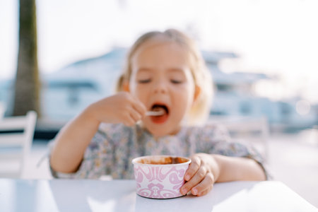 Little girl eats ice cream with a spoon, holding a cup with her hand. Blurの写真素材