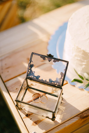 Pair of wedding rings lies in a glass box next to a wedding cake on a wooden box