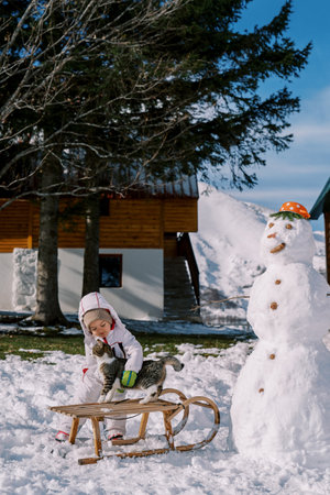 Little girl strokes a striped cat sitting on a sleigh near a snowman in the yardの写真素材