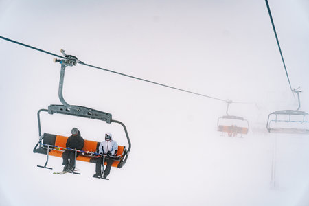 Skiers in ski suits and skis ride on a chairlift up the foggy mountainの写真素材