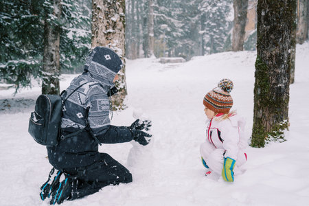 Little girl crouched and watches her mother making a snowman in the snowy forest