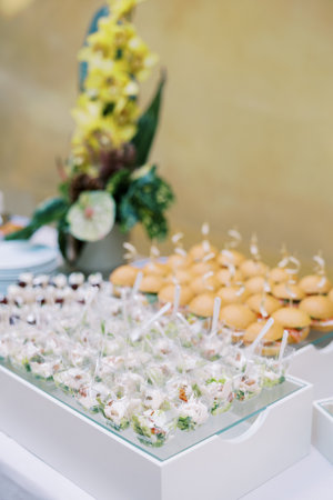 Portioned salads in cups with spoons on a tray next to mini-burgers are on the table